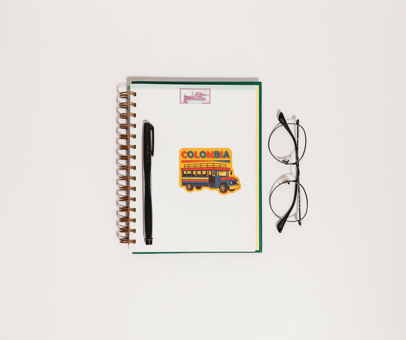 Colombia sticker with a colorful bus design on a spiral notebook, accompanied by glasses and a black pen.