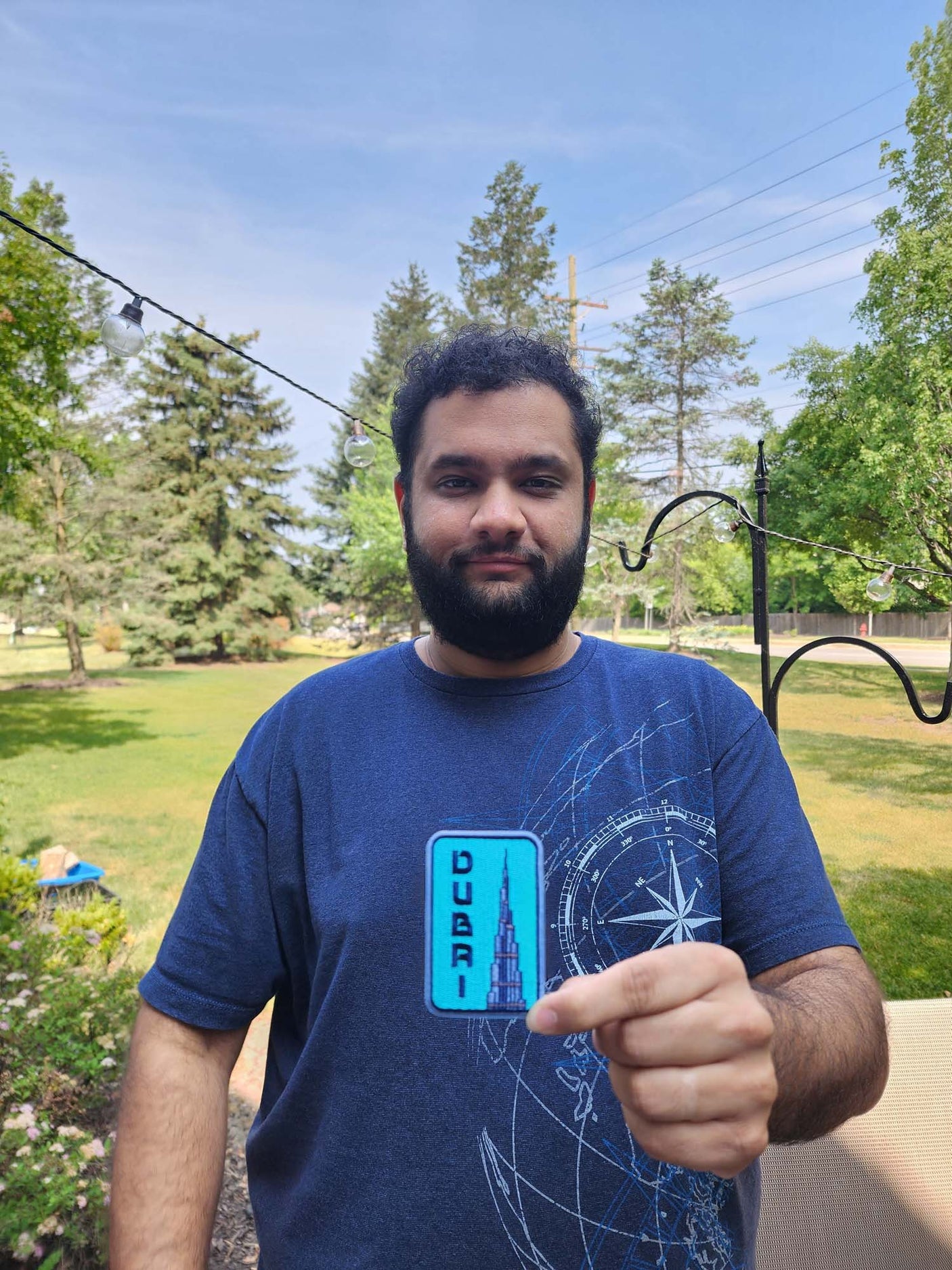 Dubai patch held by a man in a blue t-shirt, with trees and a lawn in the background.
