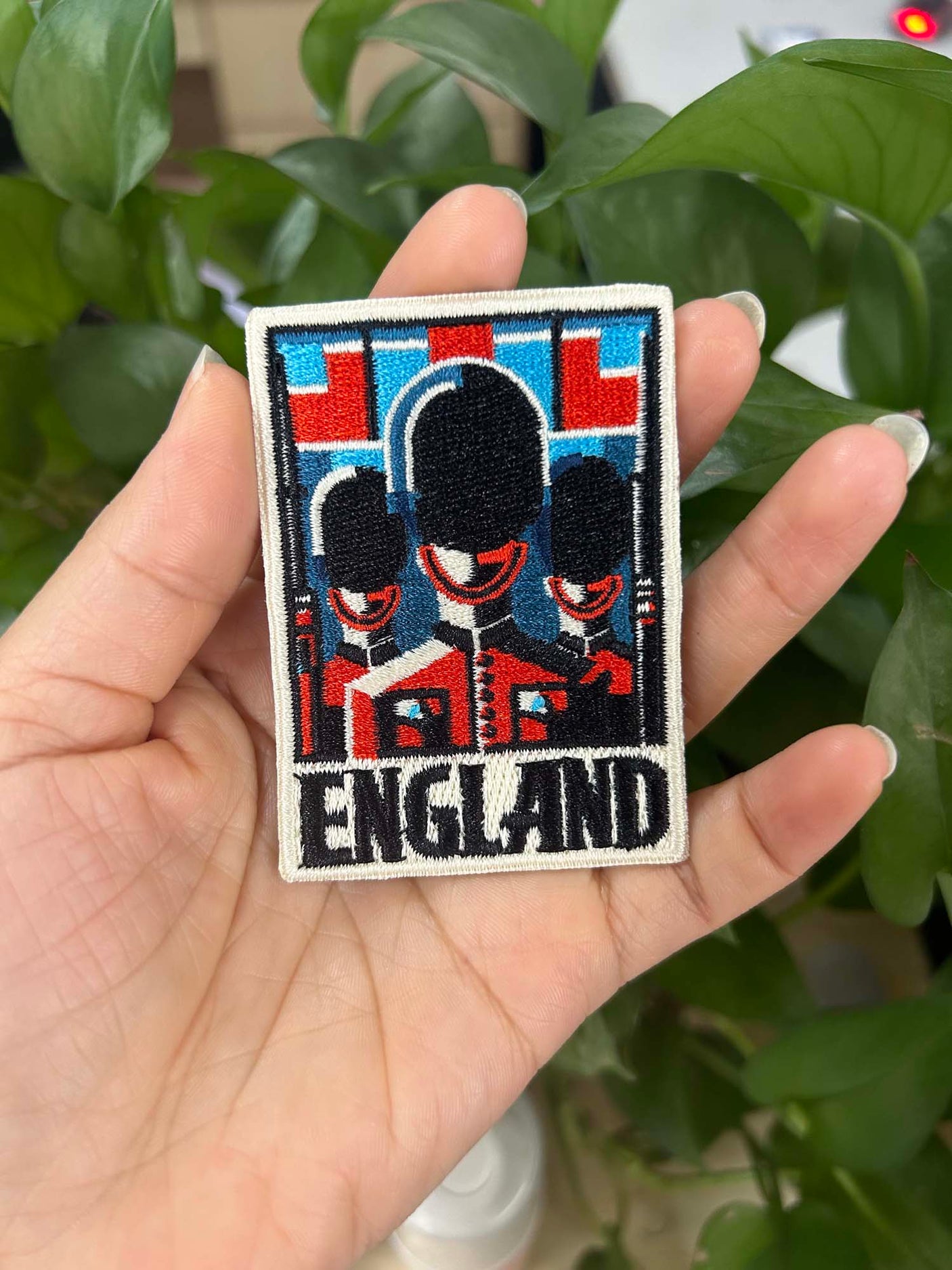 England patch featuring three guards in front of a red and blue background, held in a hand among green foliage.