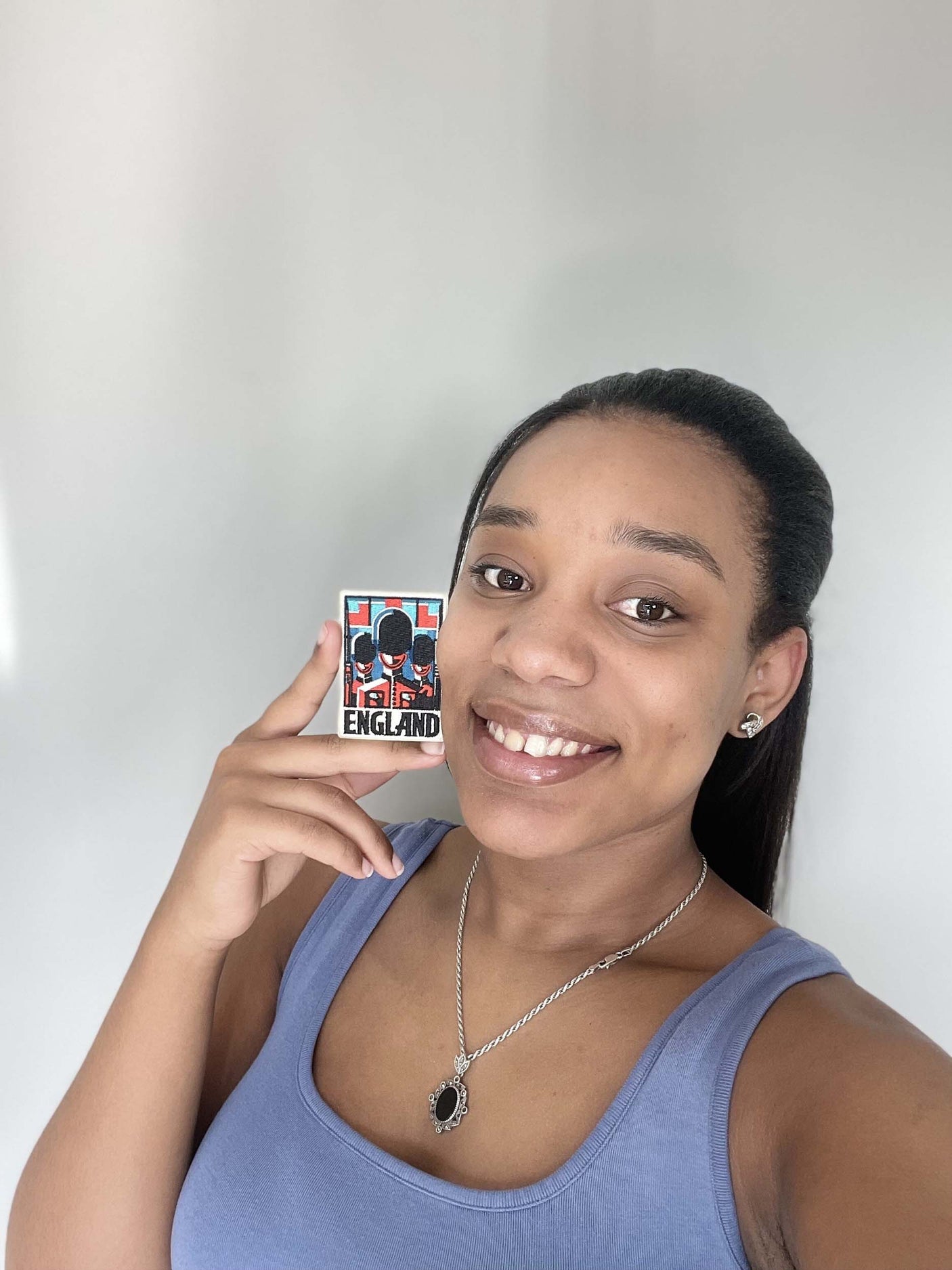 A woman with a ponytail holds an England patch, smiling and wearing a blue tank top against a light background.