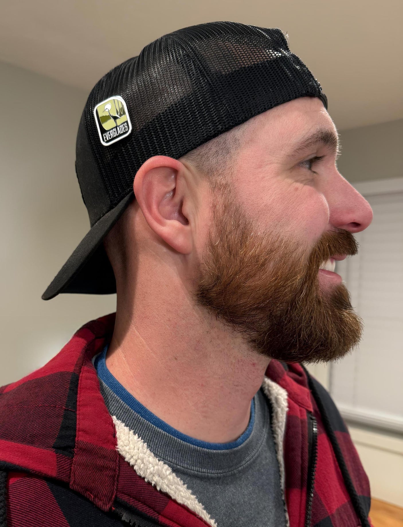 Everglades National Park enamel pin displayed on a black cap worn by a man with a beard and plaid jacket.