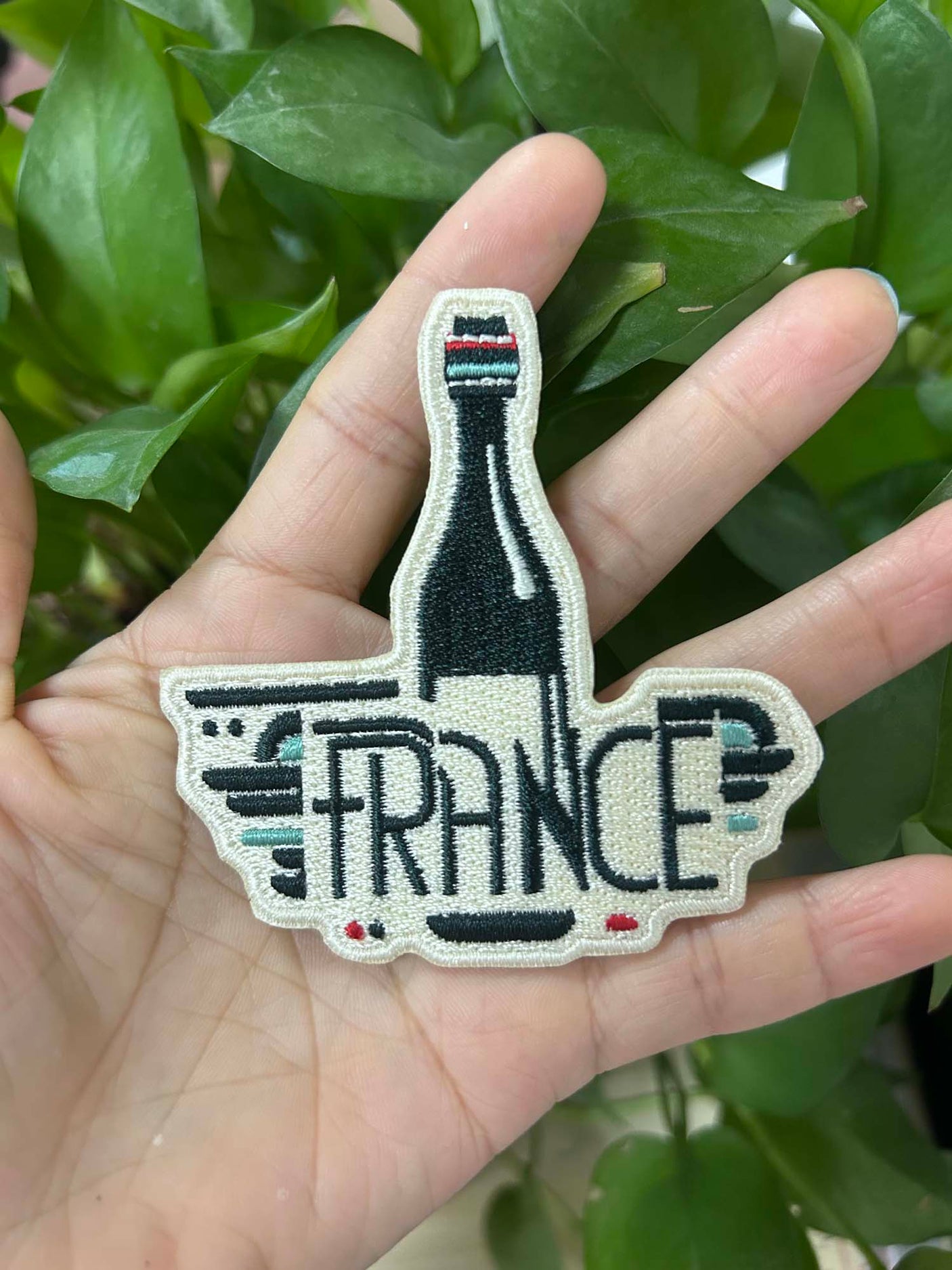 France patch featuring a bottle design with the word "FRANCE" in bold letters, held in a hand against green foliage.