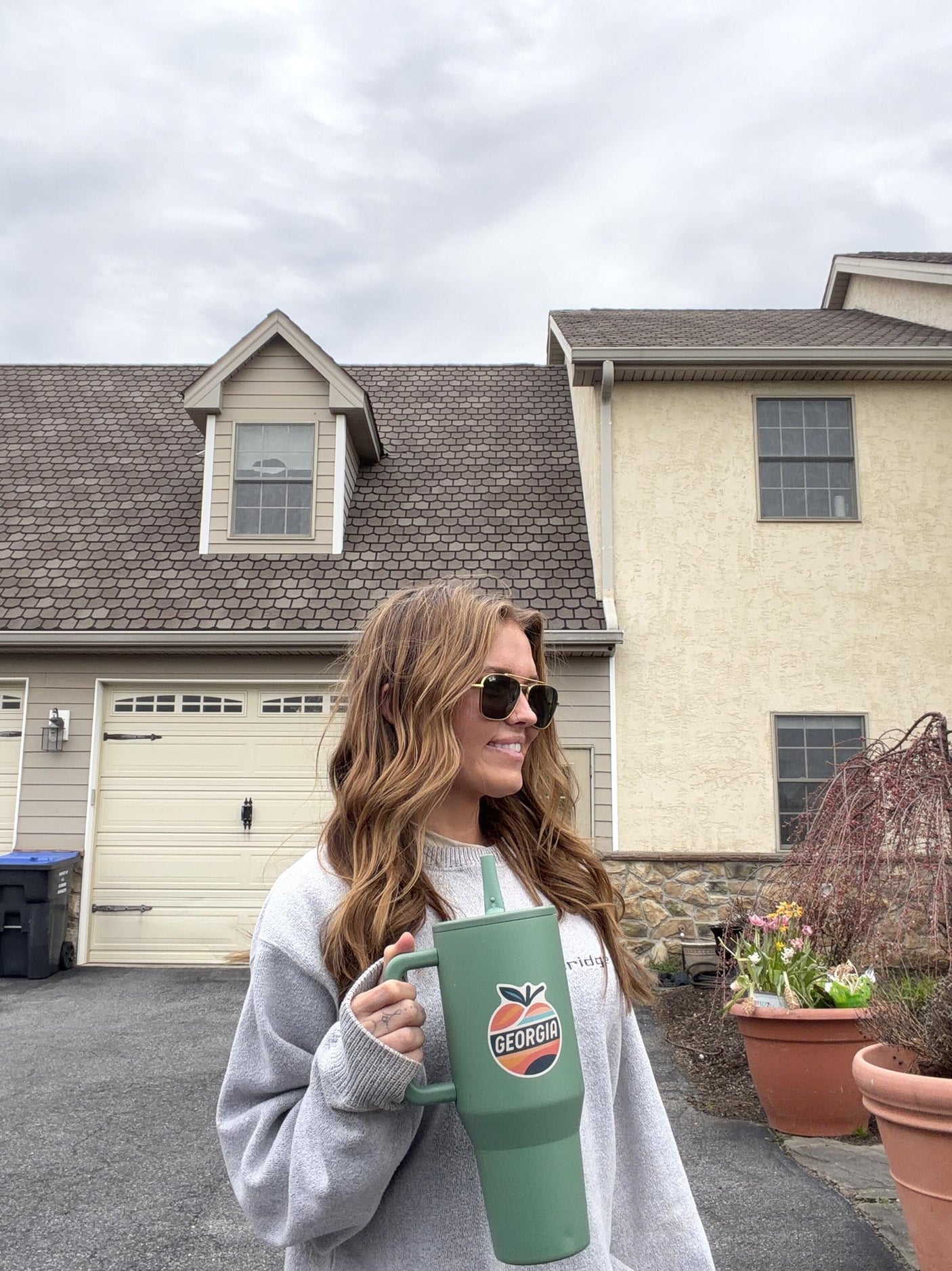 Woman wearing sunglasses holds a green mug featuring a Georgia sticker, standing in front of a house.
