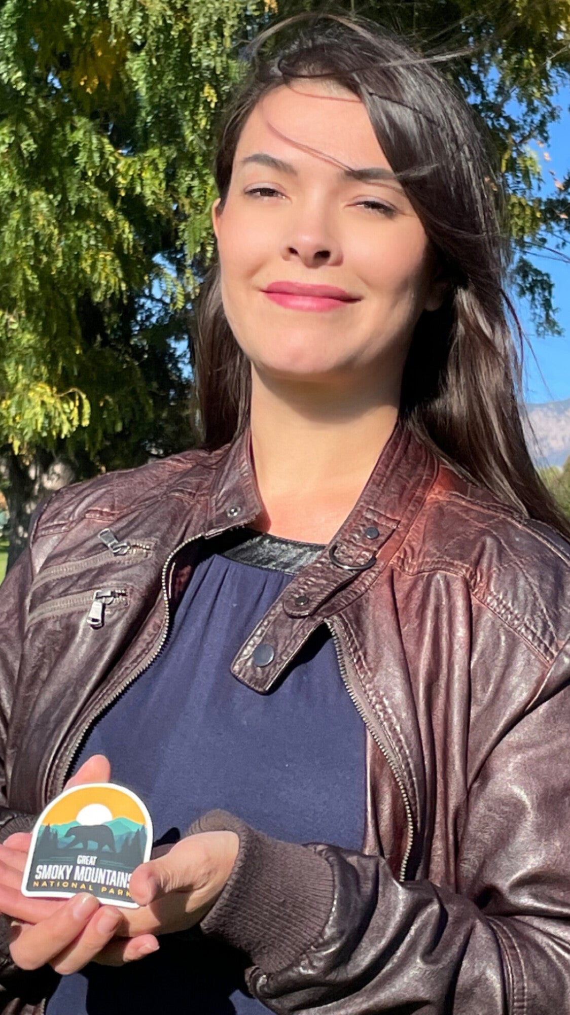 Great Smoky Mountains National Park sticker held by a woman in a brown leather jacket outdoors.