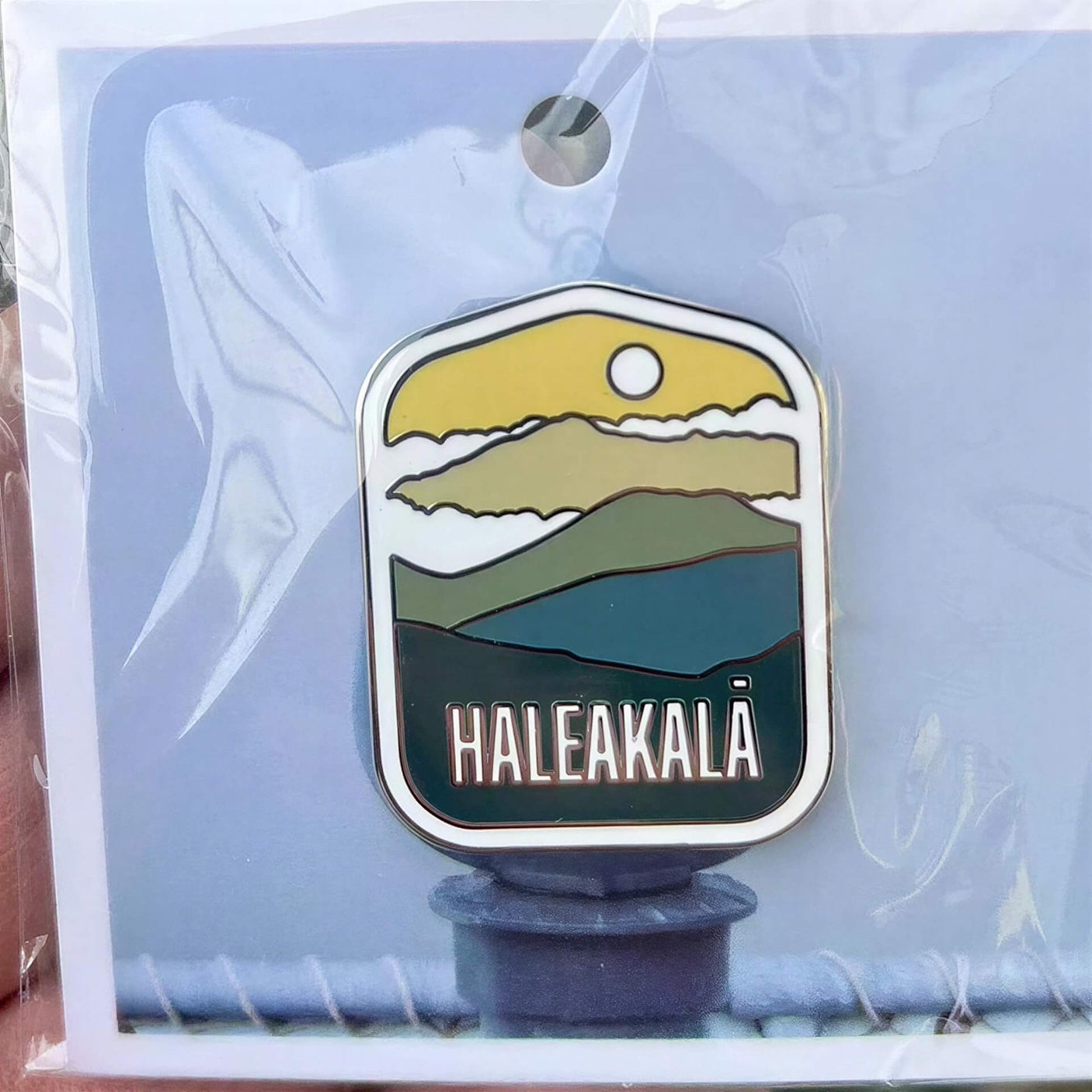 Haleakala National Park enamel pin featuring mountains, sun, and blue-green colors in a protective plastic sleeve.