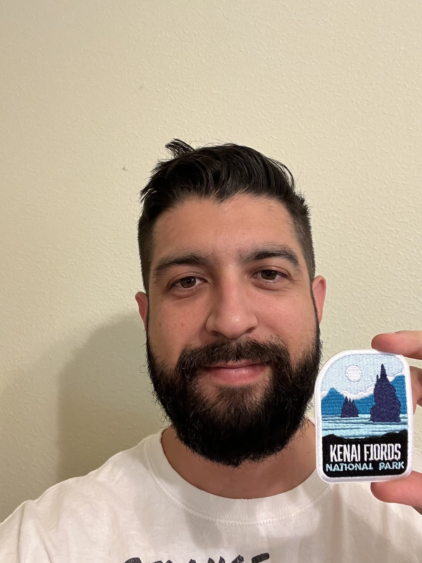 Kenai Fjords National Park patch held by a man with a beard, featuring mountains and a sun in blue and white colors.