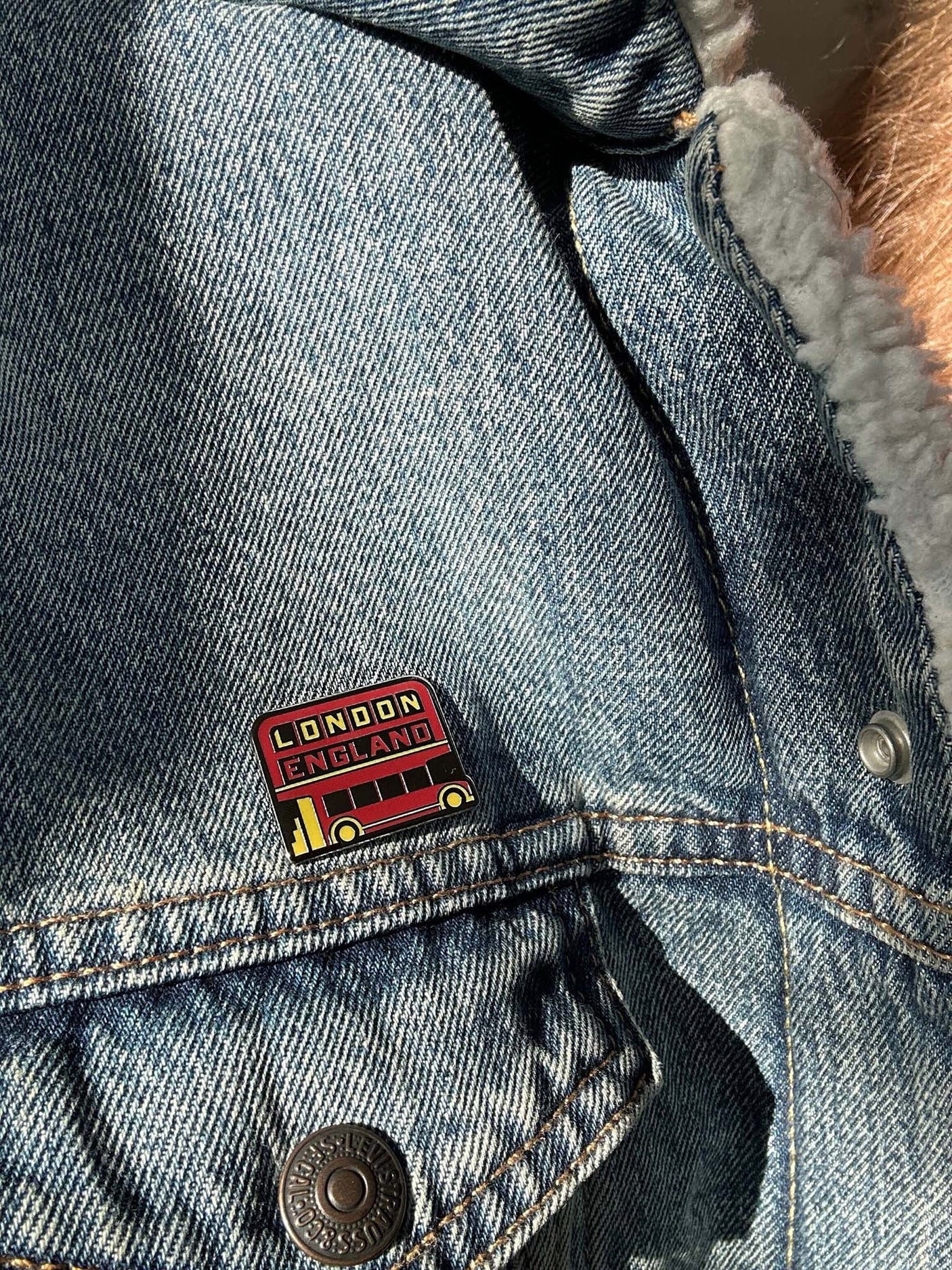 London England enamel pin featuring a red double-decker bus design on a denim jacket.