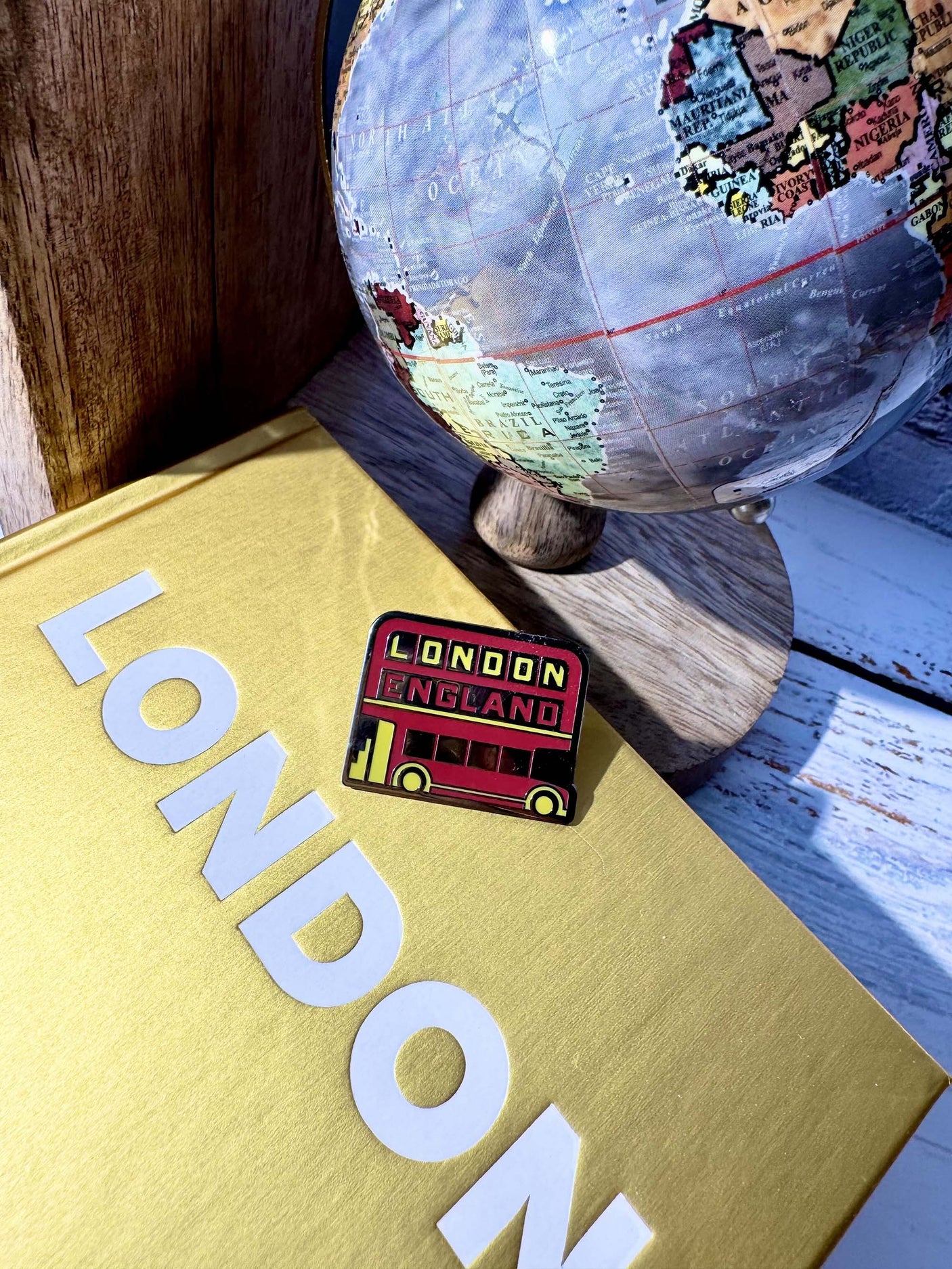 London England enamel pin featuring a red double-decker bus, placed on a gold book with "LONDON" text and a globe.
