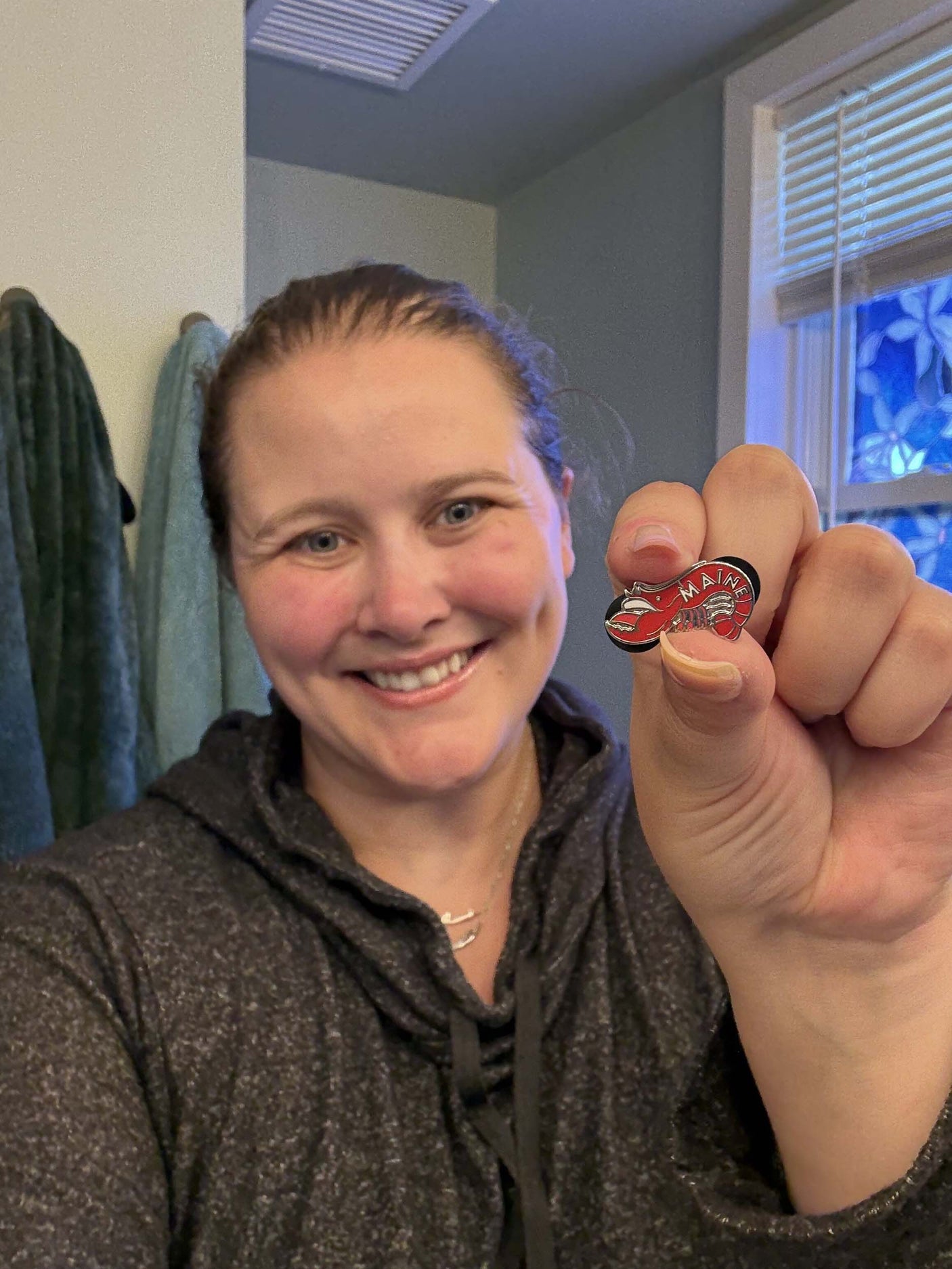 Maine enamel pin held by a smiling person, featuring a red and gold design with the word "Maine.