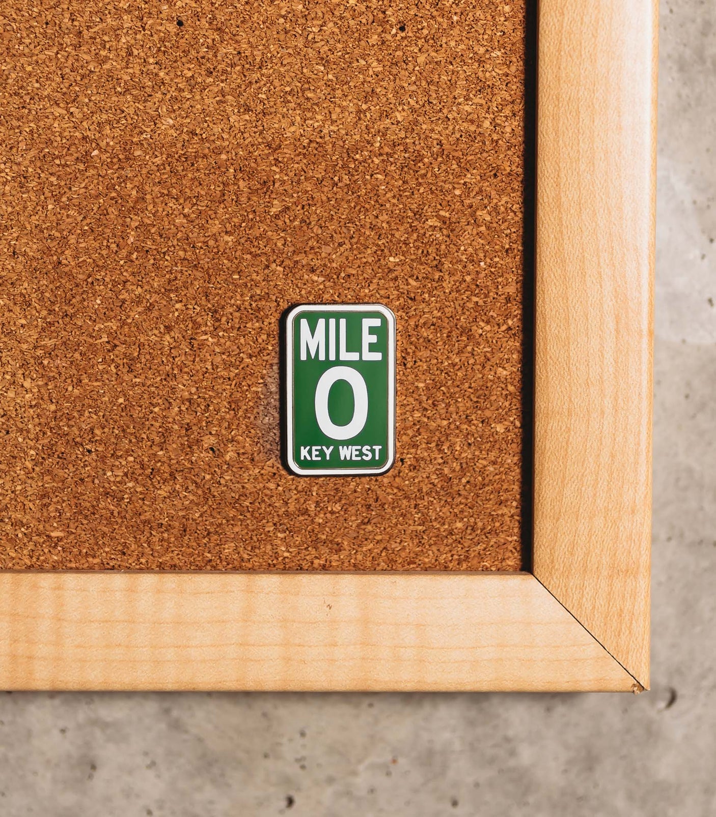 Key West Mile 0 enamel pin with a green background, white text, displayed on a corkboard with a wooden frame.