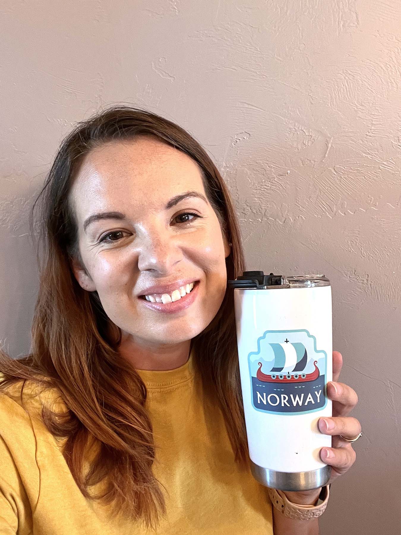 A woman holds a white tumbler featuring a Norway sticker with a ship and the word "NORWAY" in blue.