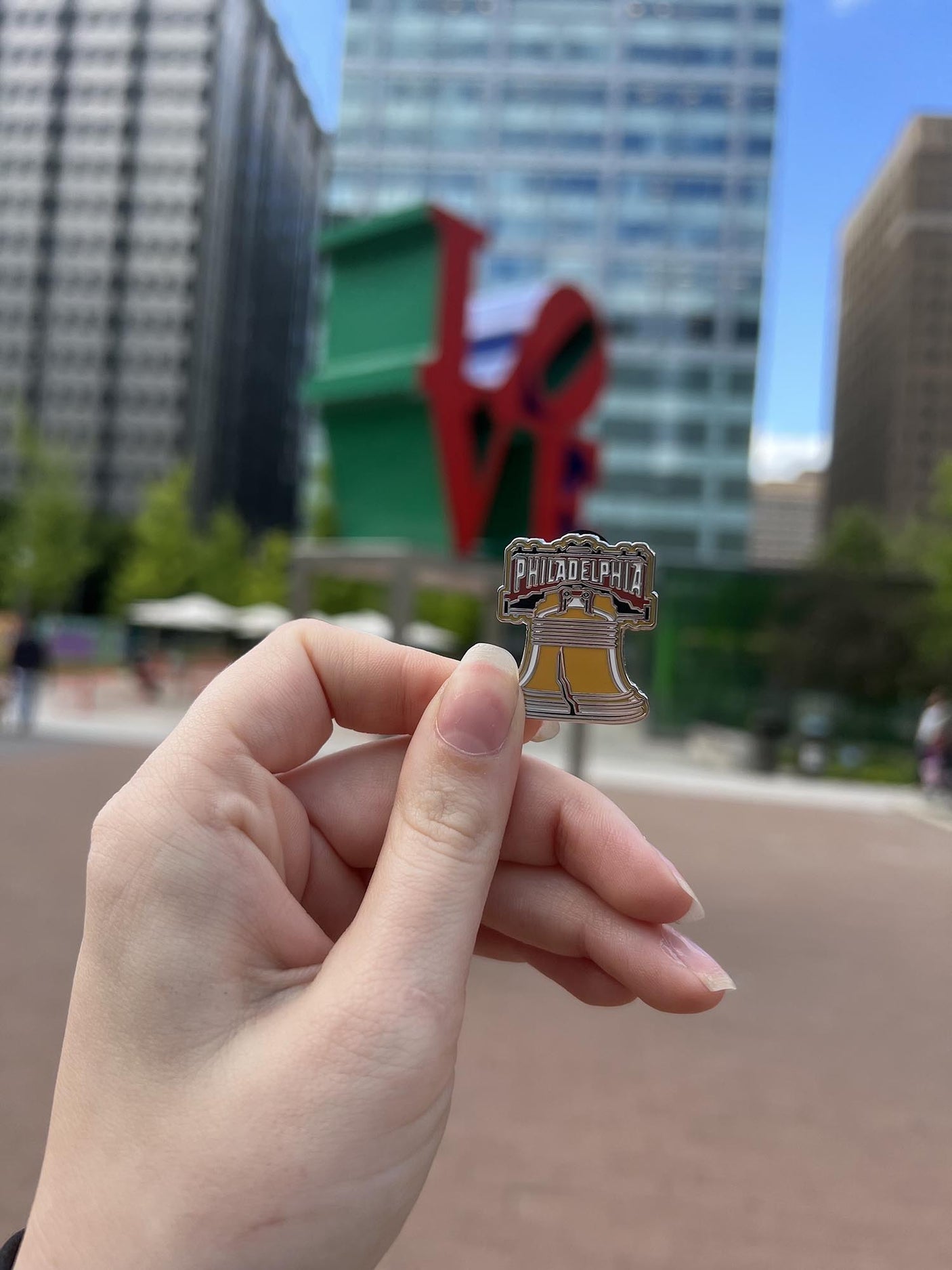 Philadelphia enamel pin featuring the Liberty Bell, held in hand with the LOVE sculpture in the background.
