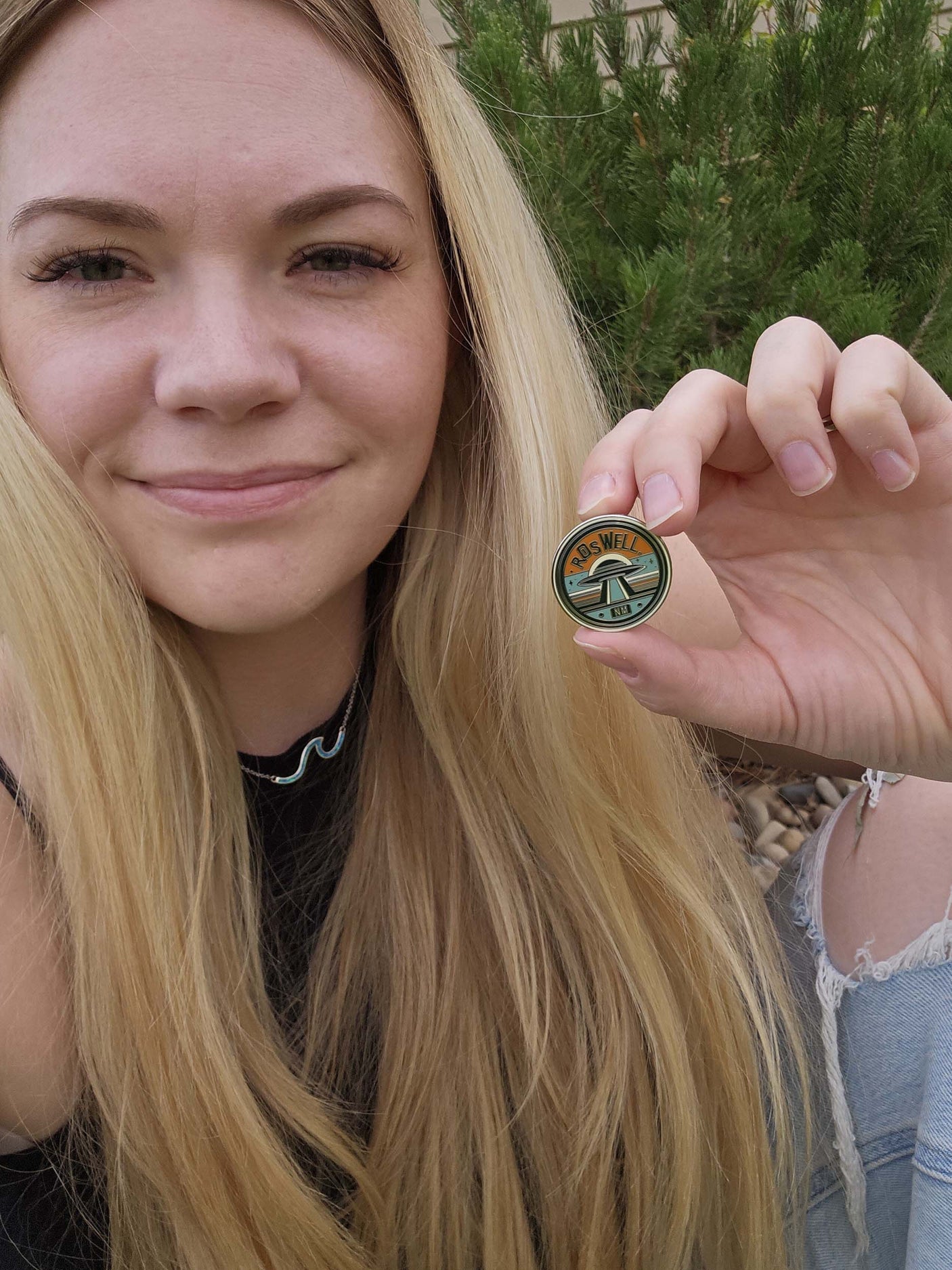 Roswell, NM enamel pin held by a person with long blonde hair, featuring a green and orange design.