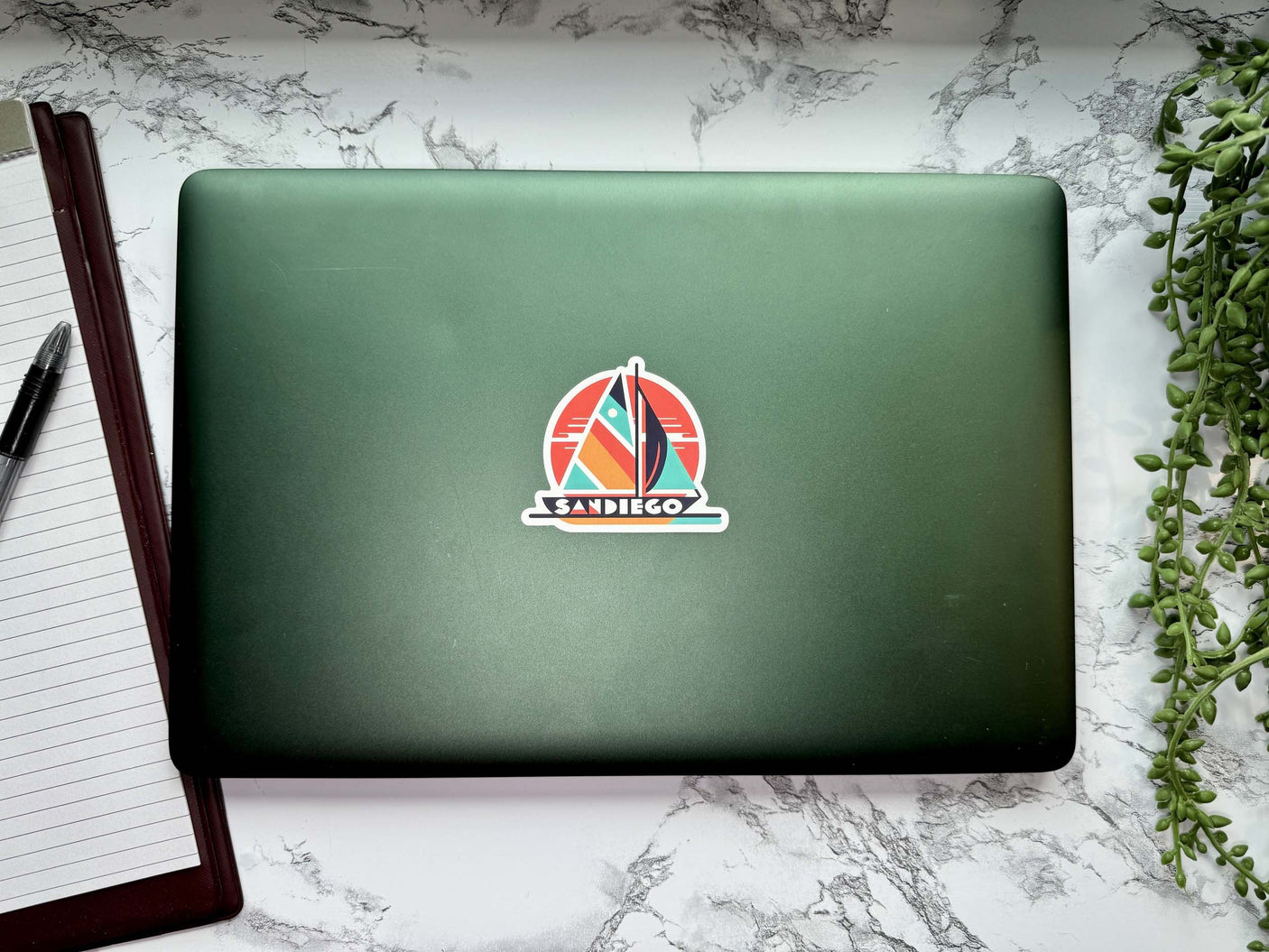 San Diego sticker featuring a sailboat design on a green laptop with a notebook and pen nearby.