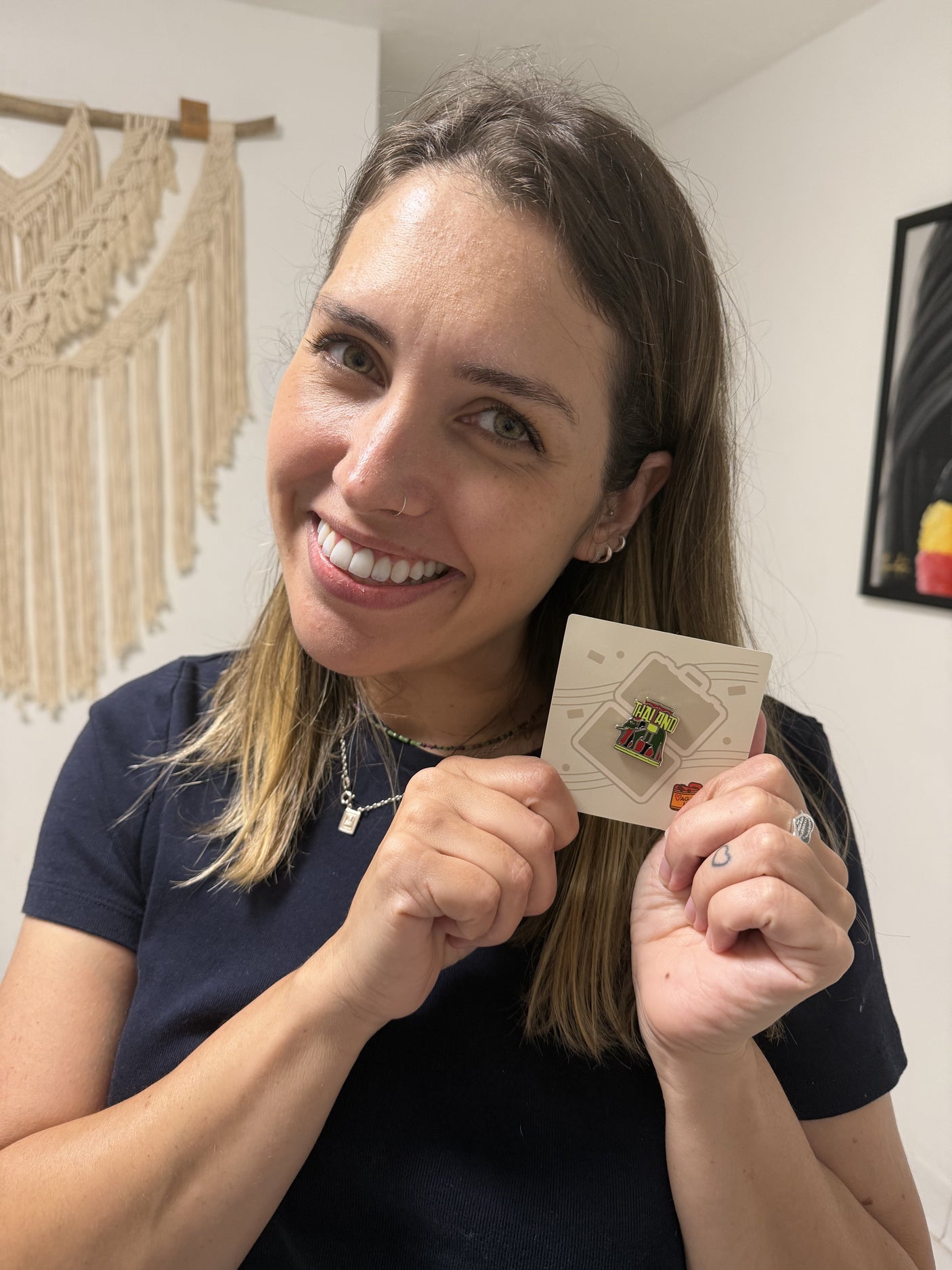 Woman holding a card with a thailand pin, smiling in a room with wall art.