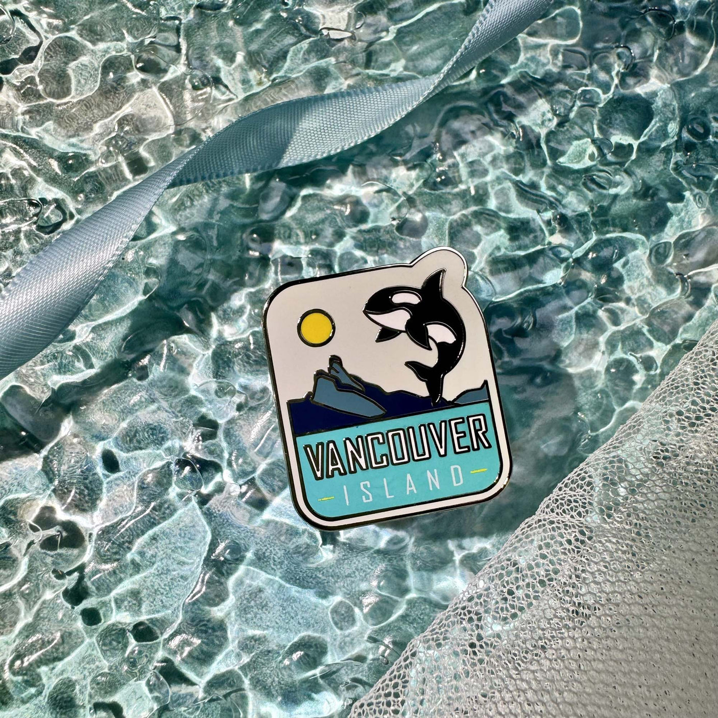 Vancouver Island enamel pin featuring a whale, mountains, sun, and blue background on a textured surface.