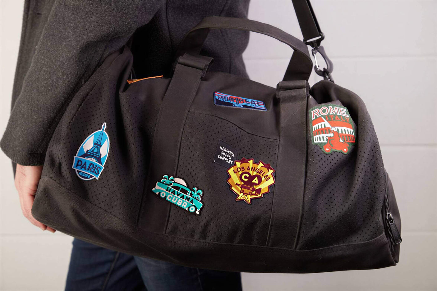 Black duffel bag with various colorful patches, including Stonehenge, Paris, and Los Angeles, displayed on a person's shou...