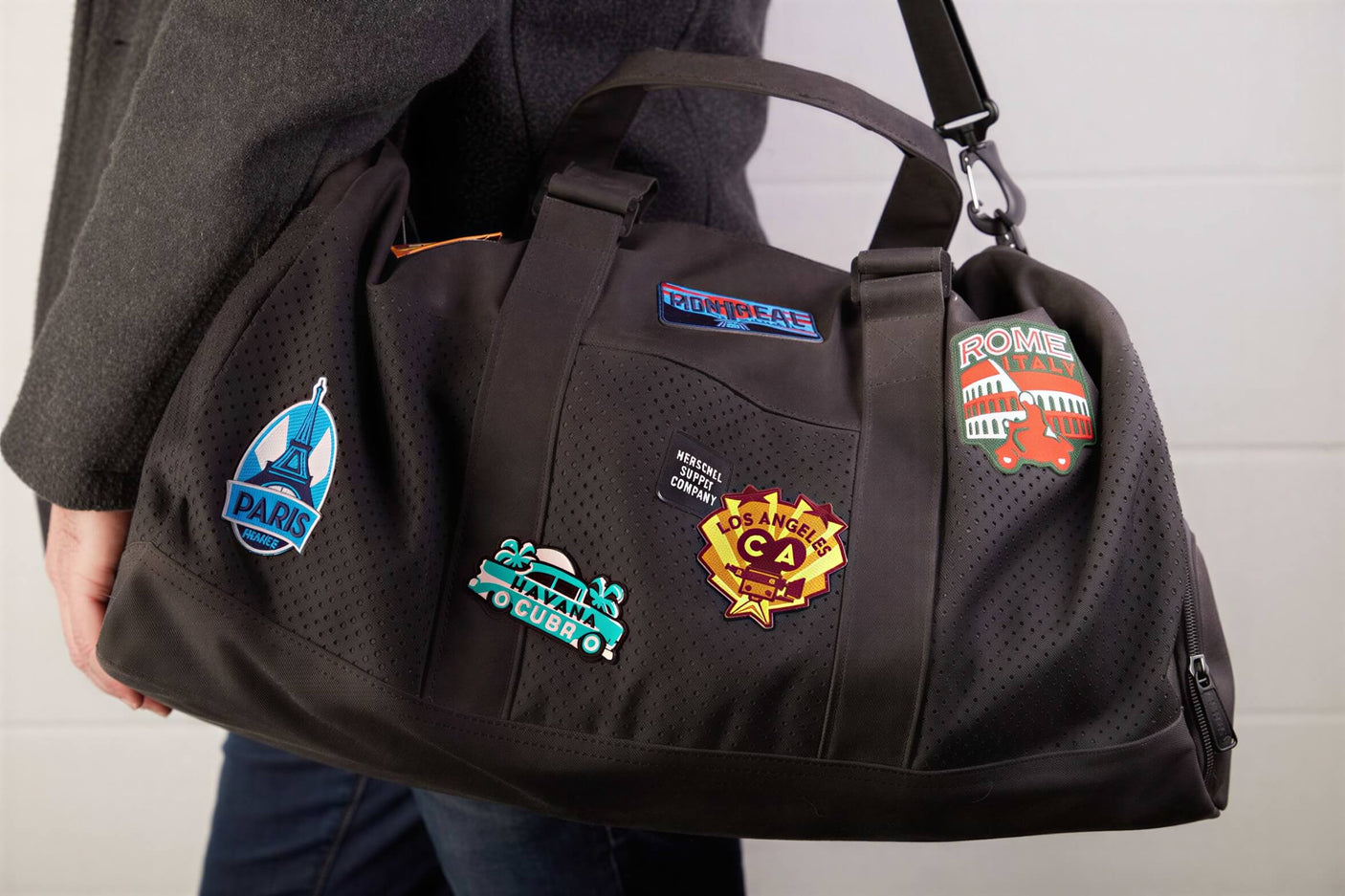 Black duffel bag with various colorful patches, including Norway, Paris, and Los Angeles, displayed on a person’s shoulder.