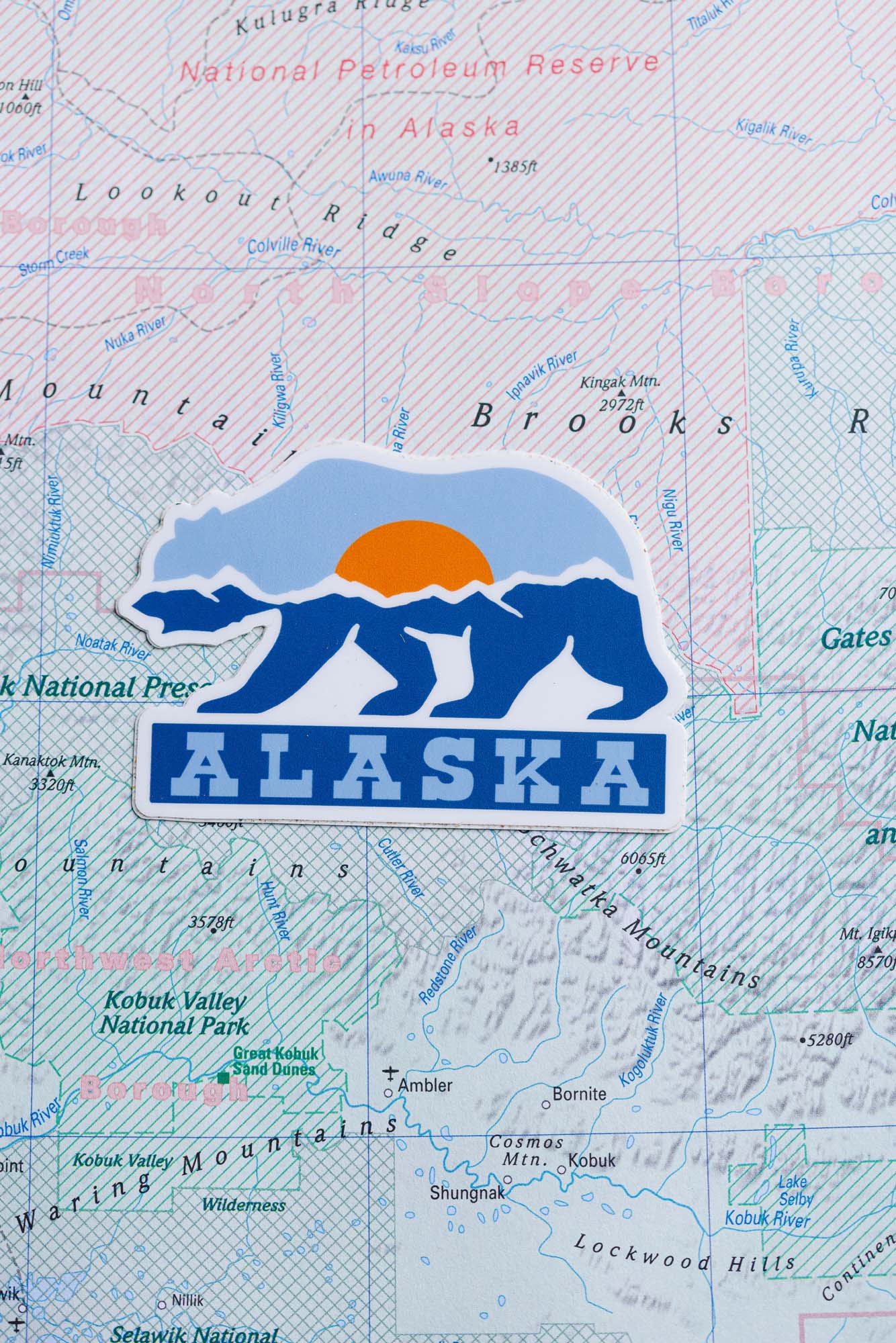 Alaska sticker featuring a bear silhouette and sun, placed on a detailed map of Alaska.