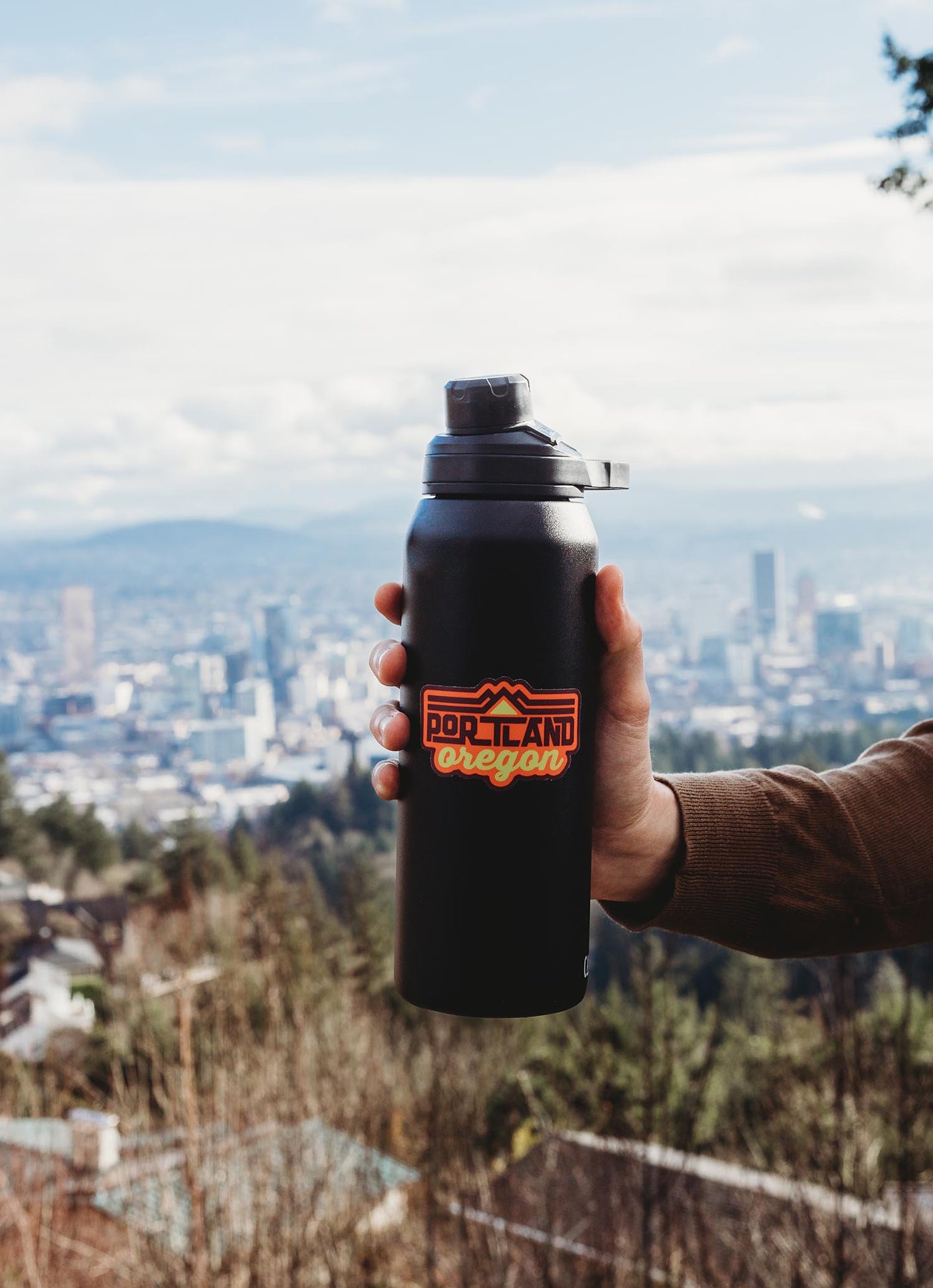 Portland Oregon sticker on a black water bottle held against a city skyline backdrop.