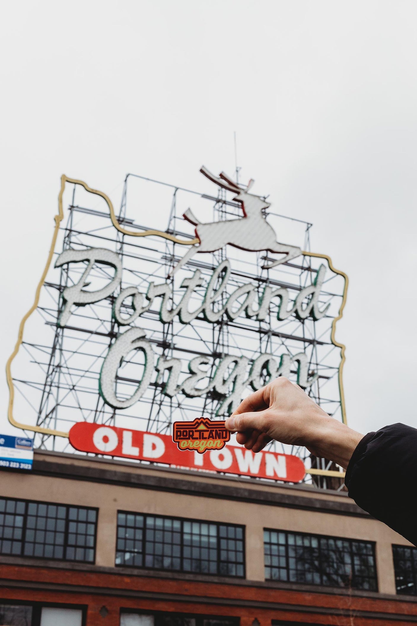 Portland Oregon sticker held in front of the iconic neon sign in Old Town, showcasing red and yellow colors.