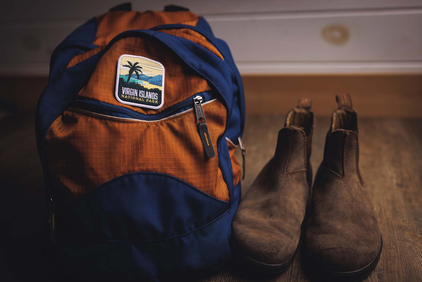 Virgin Islands National Park patch on an orange and blue backpack, with brown shoes beside it on a wooden surface.