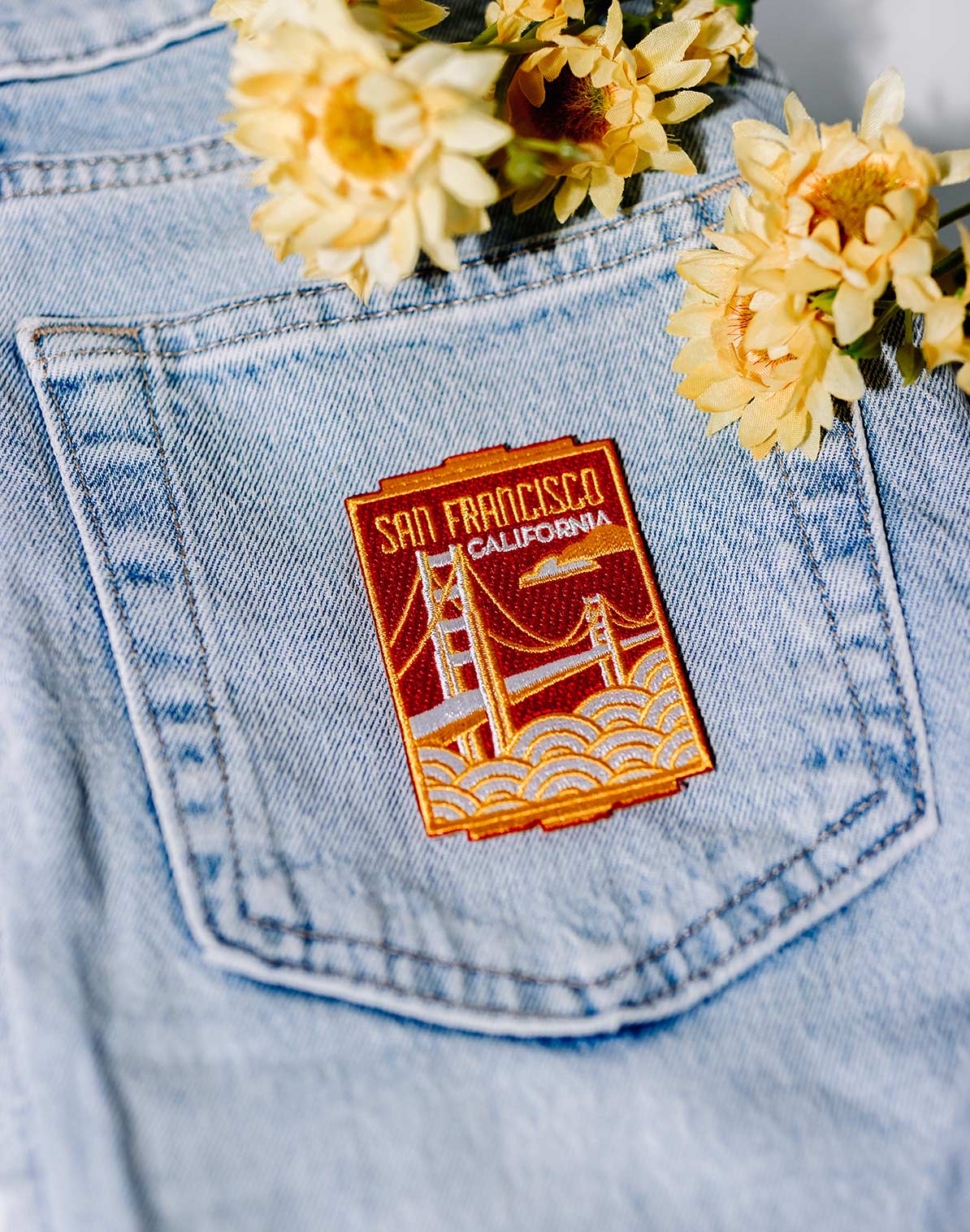 Prince Edward Island patch in orange and yellow, sewn onto a light blue denim fabric with flowers nearby.