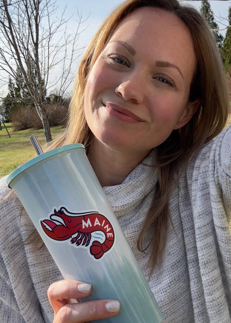 Maine sticker featuring a lobster design on a light blue tumbler held by a person outdoors.
