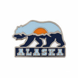 Alaska enamel pin featuring a bear silhouette, mountains, and a sunset in blue, orange, and white colors.