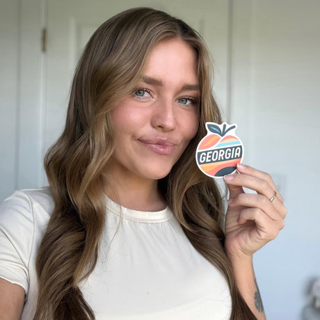 Woman holding a sticker with a peach and 'Georgia' text against a neutral background