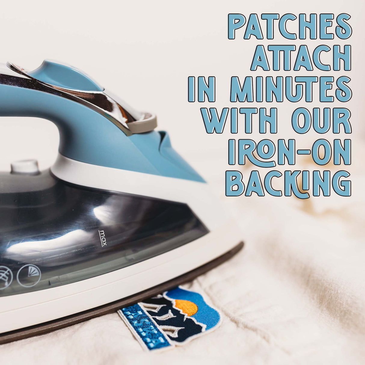 Iron-on patch with blue and white design, featuring a mountain and sun, next to a blue iron on a beige fabric.