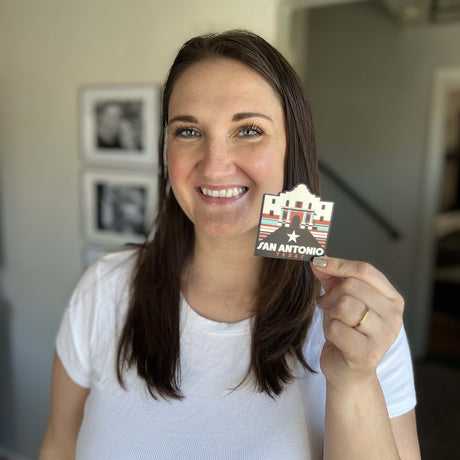 Woman holding a San Antonio sticker featuring the Alamo and red, white, and blue colors.