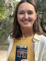 Seattle sticker on a dark tumbler held by a woman in a yellow top and white shirt, surrounded by greenery.