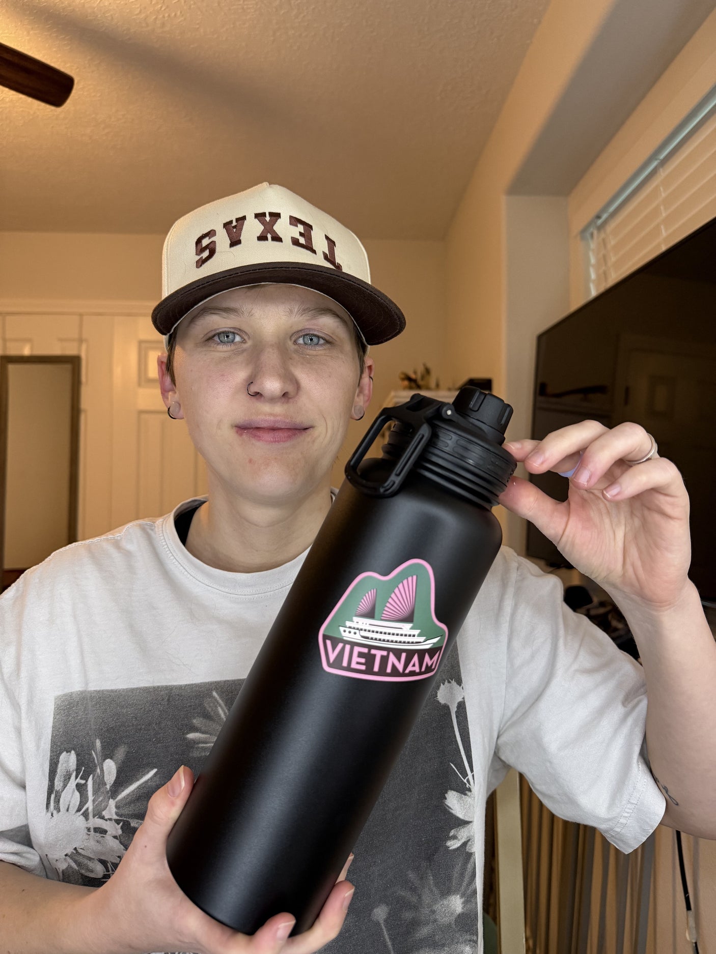 Black water bottle featuring a pink and green Vietnam sticker, held by a person wearing a beige cap and gray shirt.