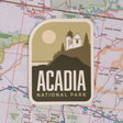 Acadia National Park sticker featuring a lighthouse and sun on a map background.